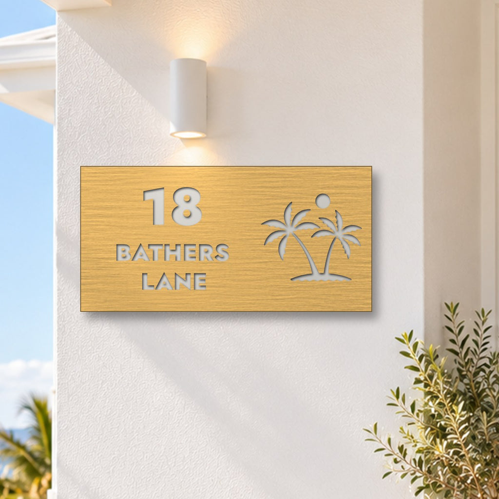 Brushed gold house number and street name sign with a brushed metal look, The Yaroomba, reading 18 Bathers Lane in white lettering, with palm trees and a sun, on a white rendered wall under a wall light, with the ocean behind.