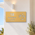 Brushed gold house number and street name sign with a brushed metal look, The Yaroomba, reading 18 Bathers Lane in white lettering, with palm trees and a sun, on a white rendered wall under a wall light, with the ocean behind.