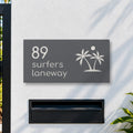 Matte charcoal house number and street name sign, The Yaroomba, reading 89 Surfers Laneway in white lettering, with palm trees and a sun, on a white rendered fence above a black letterbox.