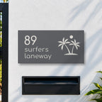 Matte charcoal house number and street name sign, The Yaroomba, reading 89 Surfers Laneway in white lettering, with palm trees and a sun, on a white rendered fence above a black letterbox.