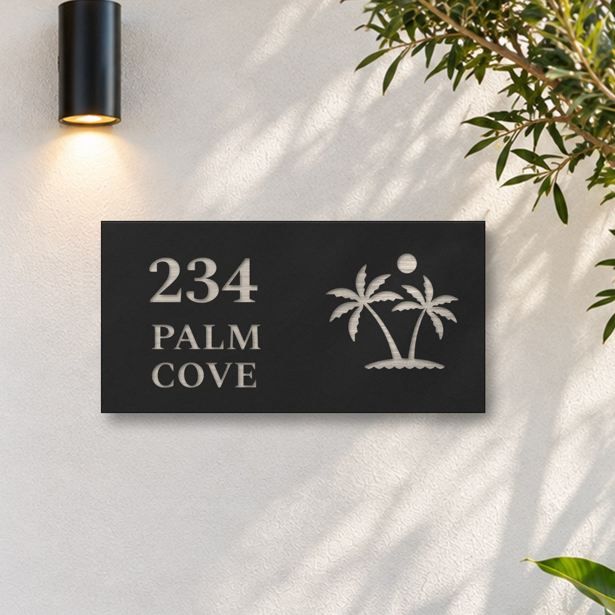 Brushed silver house number and street name sign with a brushed metal look, The Yaroomba, black face reading 234 Palm Cove in silver lettering, with palm trees and a sun, on a white rendered wall under a black wall light, beside an olive tree.