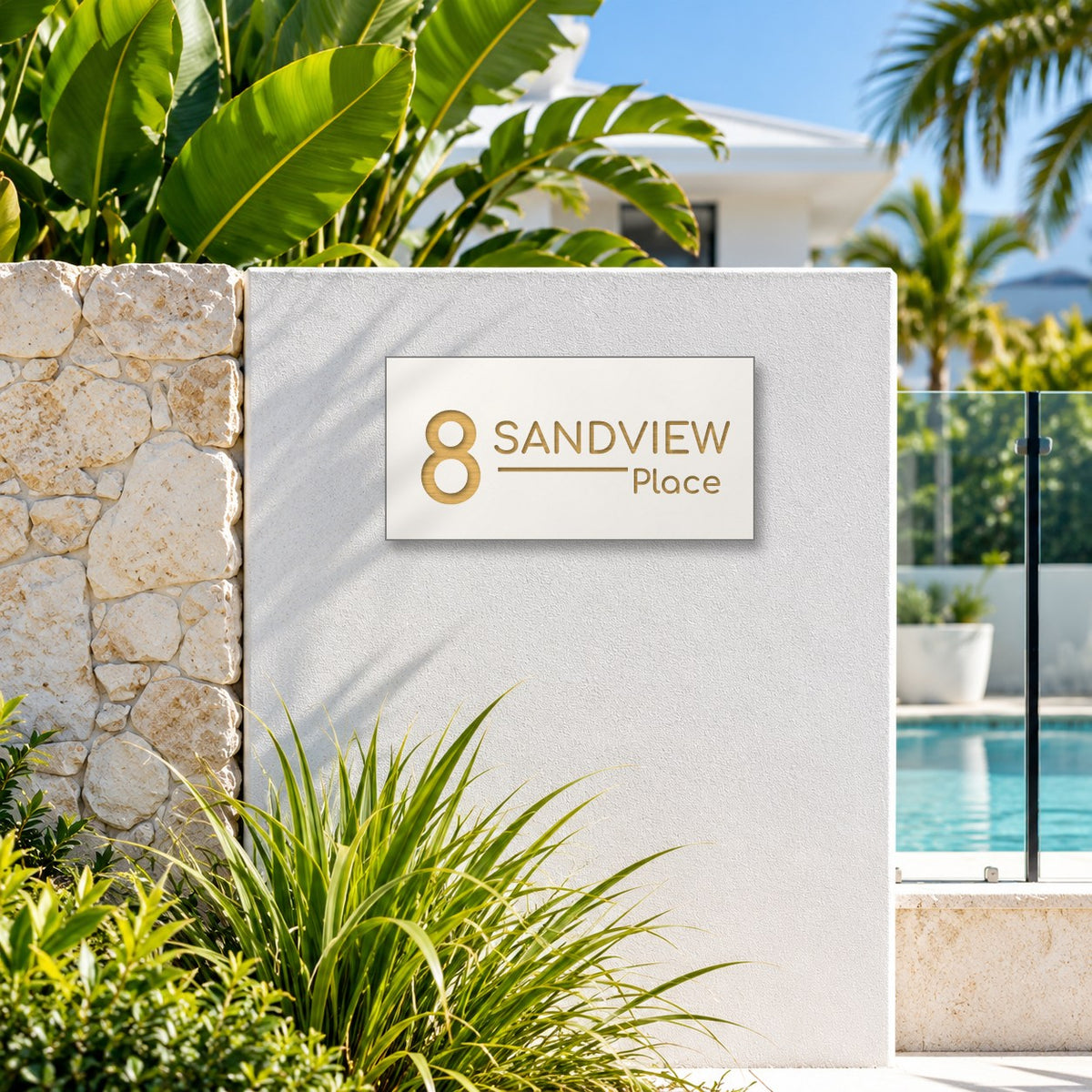 Brushed gold house number and street name sign with a brushed metal look, The Alexandra, white face reading 8 Sandview Place in gold lettering, mounted on a white rendered wall beside a stone pillar and pool fence.