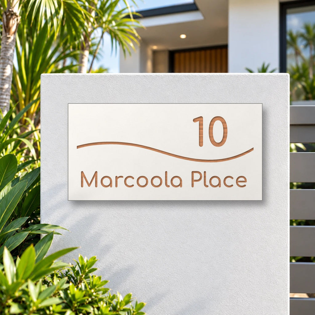 Brushed copper house number and street name sign with a brushed metal look, The Bokarina, white face reading 10 Marcoola Place in copper lettering, with a wave line, mounted on a white rendered front pillar.