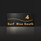 Brushed gold house number and street name sign with a brushed metal look, The Bokarina, black face reading 4 Surf Rise South in gold lettering, with a wave line.