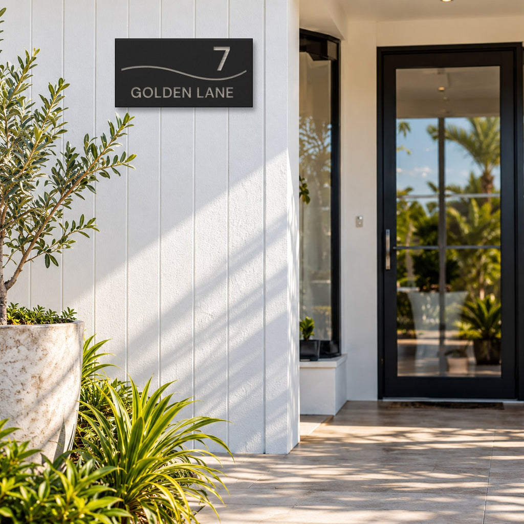 Brushed silver house number and street name sign with a brushed metal look, The Bokarina, black face reading 7 Golden Lane in silver lettering, with a wave line, mounted on white cladding beside a glass front door.