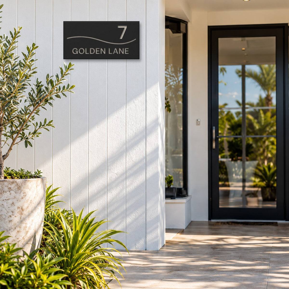 Brushed silver house number and street name sign with a brushed metal look, The Bokarina, black face reading 7 Golden Lane in silver lettering, with a wave line, mounted on white cladding beside a glass front door.