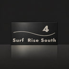 Brushed silver house number and street name sign with a brushed metal look, The Bokarina, black face reading 4 Surf Rise South in silver lettering, with a wave line.