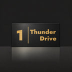 Brushed gold house number and street name sign with a brushed metal look, The Coolum, black face reading 1 Thunder Drive in gold lettering, with a dividing line.