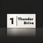 Matte white house number and street name sign, The Coolum, reading 1 Thunder Drive in black lettering, with a dividing line.
