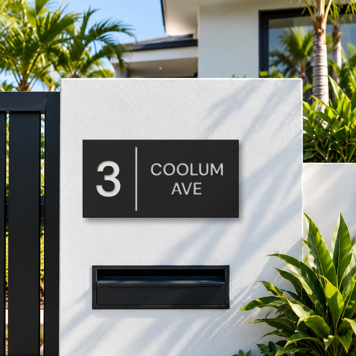Matte black house number and street name sign, The Coolum, reading 3 Coolum Ave in white lettering, with a dividing line, mounted on a white rendered fence above a black letterbox.