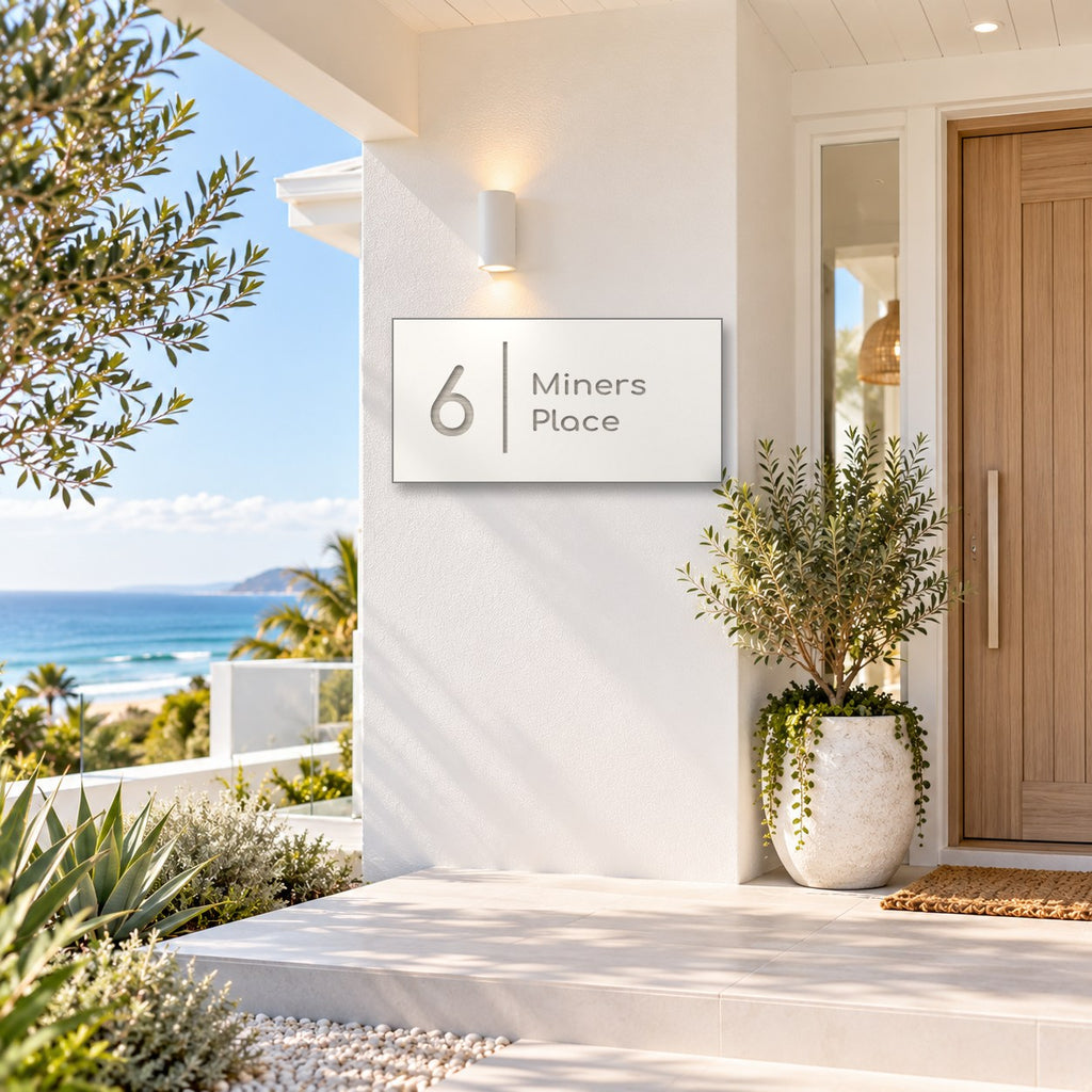 Brushed silver house number and street name sign with a brushed metal look, The Coolum, white face reading 6 Miners Place in silver lettering, with a dividing line, mounted on a white rendered wall beside a timber front door, with the ocean behind.