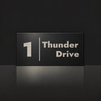 Brushed silver house number and street name sign with a brushed metal look, The Coolum, black face reading 1 Thunder Drive in silver lettering, with a dividing line.