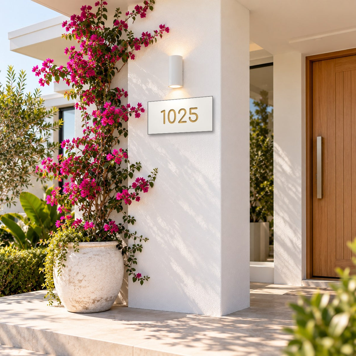 Brushed gold house number sign with a brushed metal look, The Kawana, white face with the number 1025 in gold lettering, mounted on a white rendered column beside a bougainvillea.