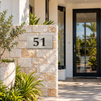 Brushed silver house number sign with a brushed metal look, The Kawana, with the number 51 in black lettering, mounted on a sandstone block pillar beside a glass front door.