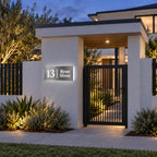 Brushed silver illuminated house number and street name sign with a brushed metal look, The Coolum, mounted on a white rendered entry wall beside the gate of a modern home at dusk, 13 Ryan Street lit cool white.