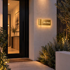Brushed gold illuminated house number sign with a brushed metal look, The Montville, mounted on a rendered wall beside a dark front door at dusk with a warm halo around it, the number spelled out as fifty three.