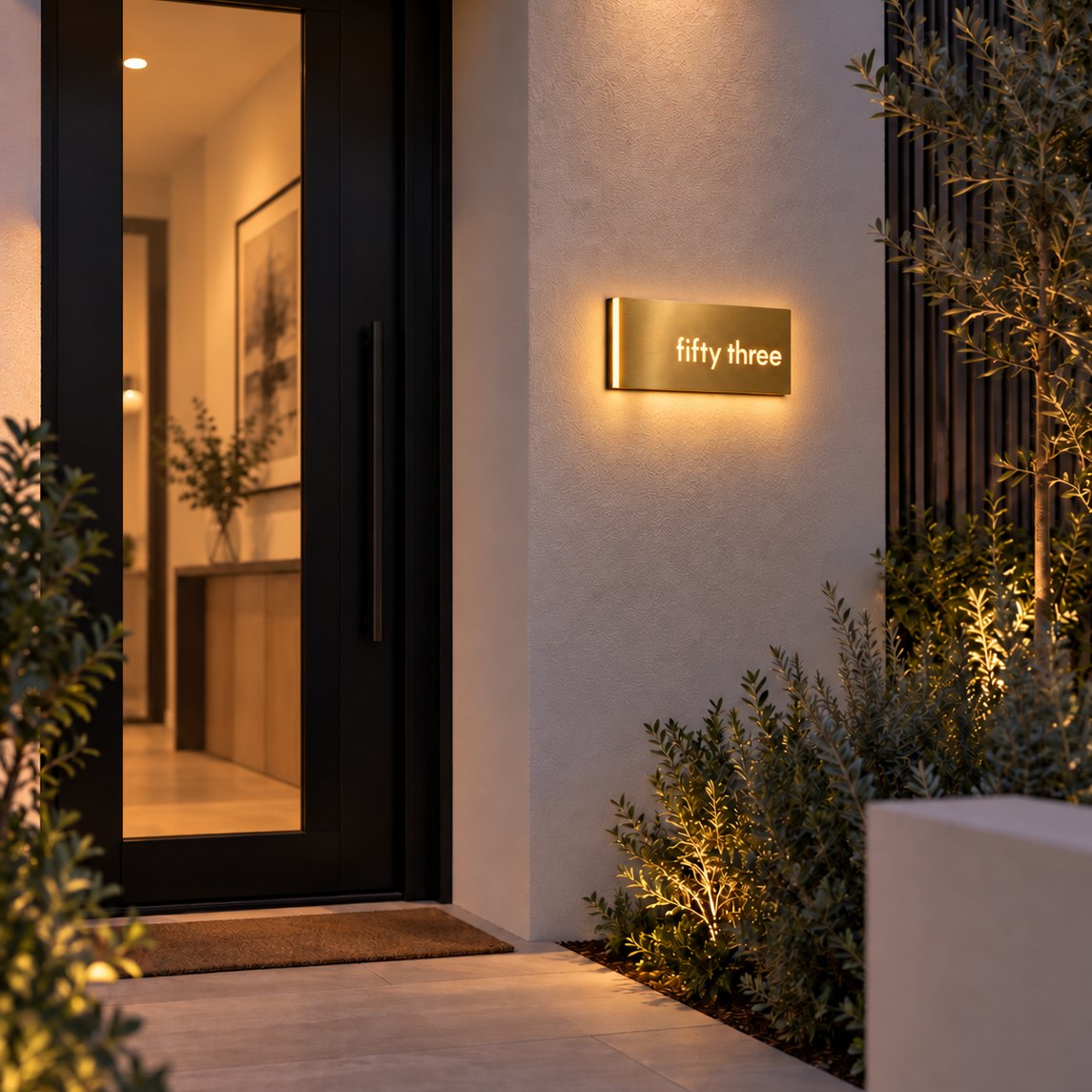 Brushed gold illuminated house number sign with a brushed metal look, The Montville, mounted on a rendered wall beside a dark front door at dusk with a warm halo around it, the number spelled out as fifty three.