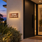 Brushed copper illuminated house number and street name sign with a brushed metal look, The Noosa, mounted on a rendered wall beside a dark front door at dusk, 2098 Pacific Highway lit warm white.