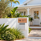 Brushed copper house number and street name sign with a brushed metal look, The Maleny, reading 16 Pelican Way in black lettering, mounted on a white fence pillar above a white letterbox, in front of a Queenslander.