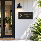 Brushed gold house number and street name sign with a brushed metal look, The Maleny, black face reading 2 Bunnytail Ave in gold lettering, mounted on white cladding beside a black front door.