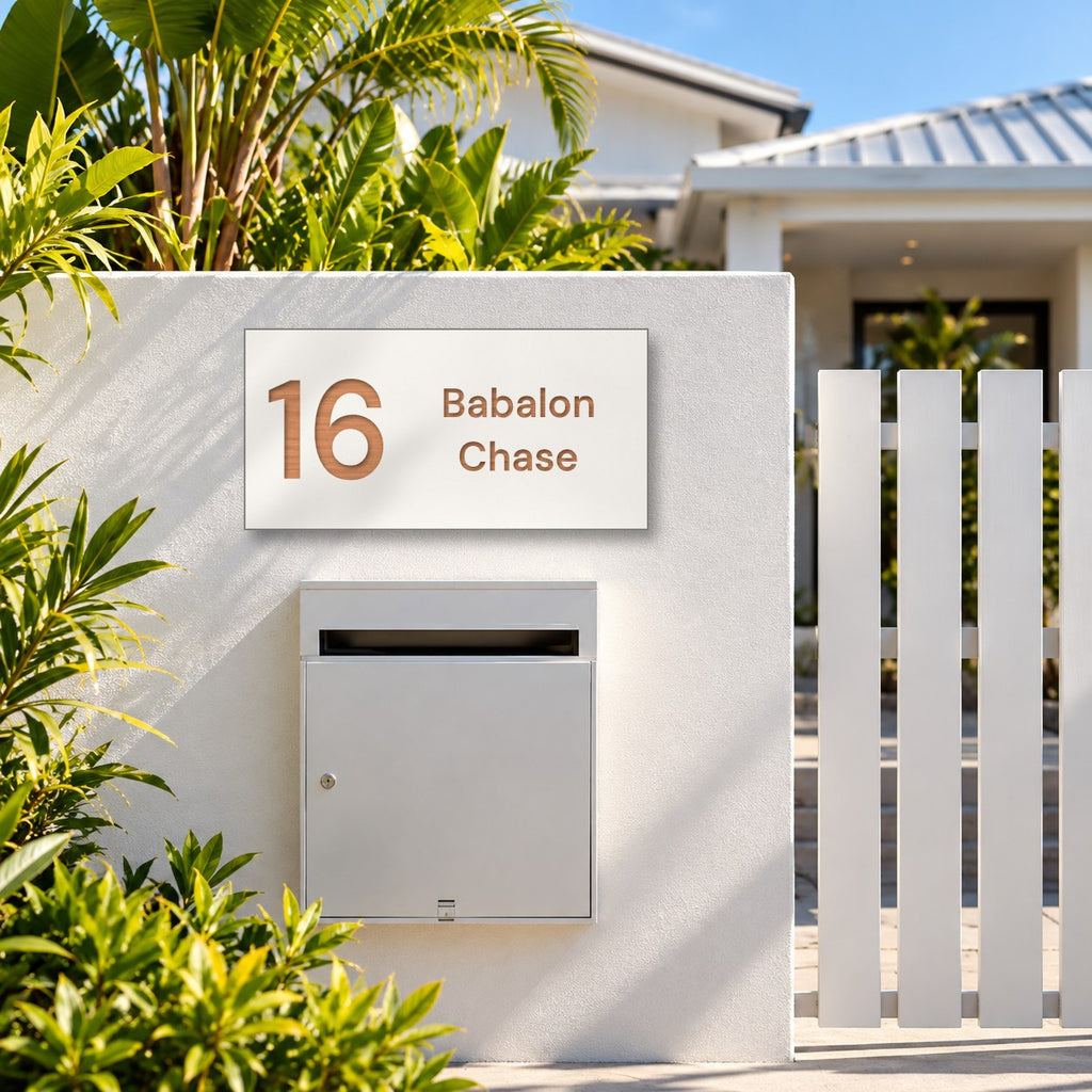 Brushed copper house number and street name sign with a brushed metal look, The Marcoola, white face reading 16 Babalon Chase in copper lettering, mounted on a white rendered fence above a silver letterbox.