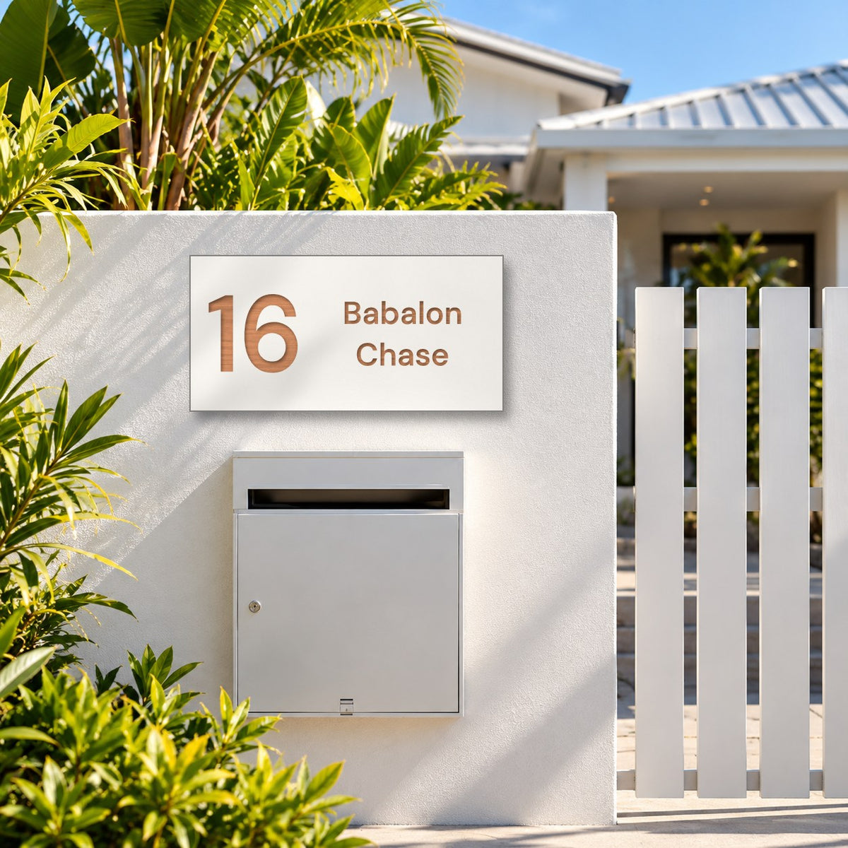 Brushed copper house number and street name sign with a brushed metal look, The Marcoola, white face reading 16 Babalon Chase in copper lettering, mounted on a white rendered fence above a silver letterbox.