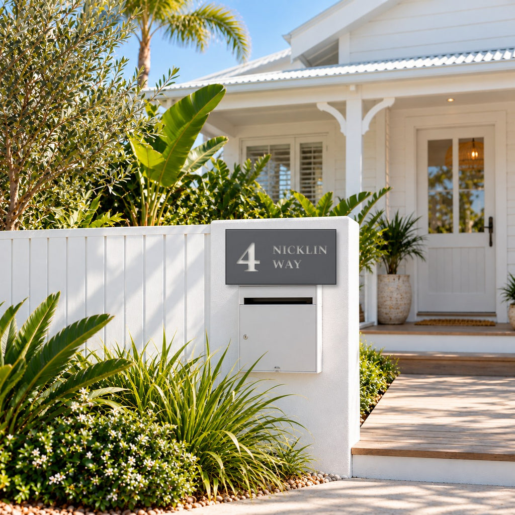 Matte charcoal house number and street name sign, The Marcoola, reading 4 Nicklin Way in white lettering, mounted on a white fence pillar above a white letterbox, in front of a Queenslander.