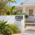 Matte charcoal house number and street name sign, The Marcoola, reading 4 Nicklin Way in white lettering, mounted on a white fence pillar above a white letterbox, in front of a Queenslander.