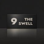 Brushed silver house number and street name sign with a brushed metal look, The Marcoola, black face reading 9 The Swell in silver lettering.