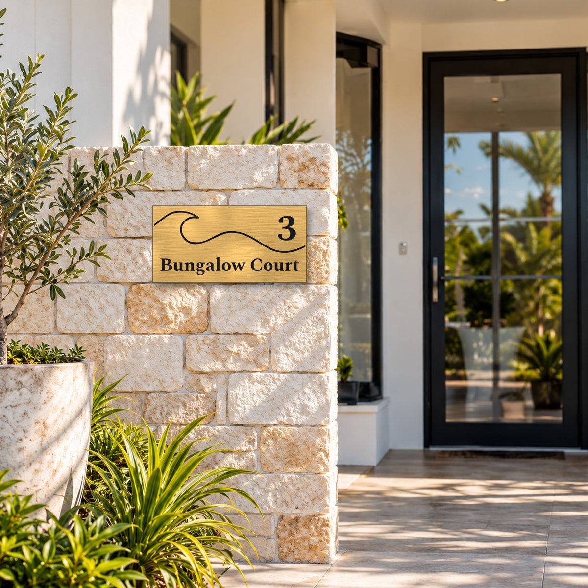 Brushed gold house number and street name sign with a brushed metal look, The Moffat, reading 3 Bungalow Court in black lettering, with a wave, mounted on a sandstone block pillar beside a glass front door.