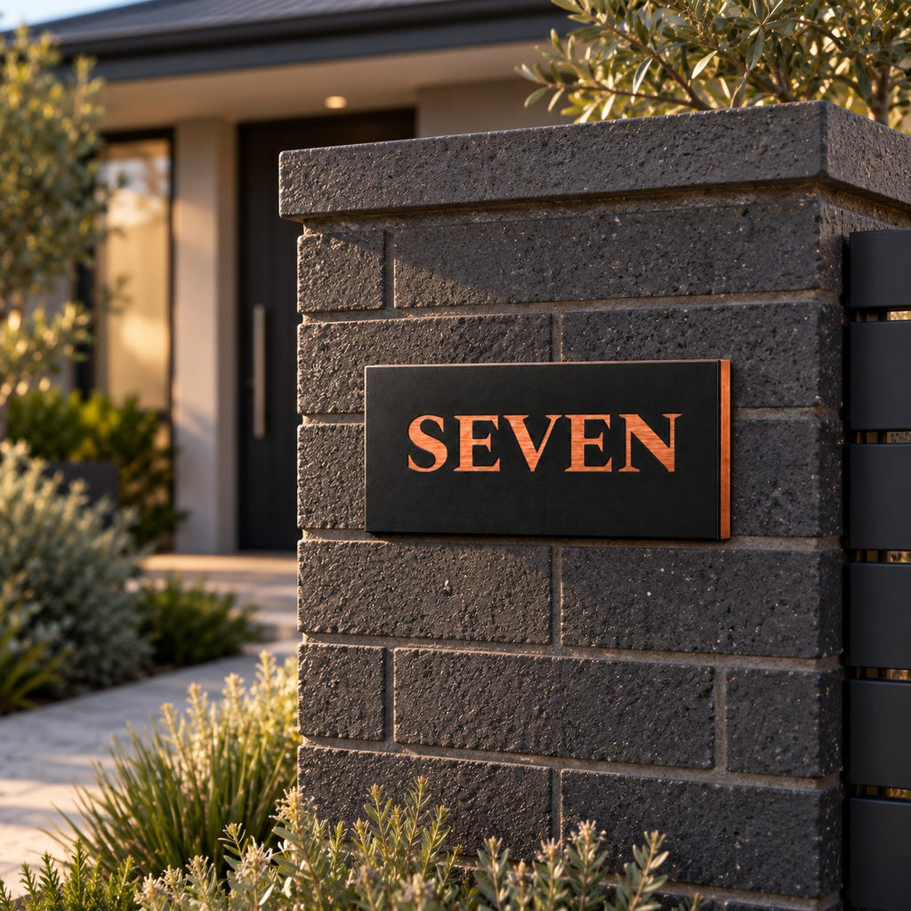 Brushed copper house number sign with a brushed metal look, The Montville, black face with the number spelled out as SEVEN in copper lettering, mounted on a charcoal block pillar beside a front path.