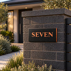 Brushed copper house number sign with a brushed metal look, The Montville, black face with the number spelled out as SEVEN in copper lettering, mounted on a charcoal block pillar beside a front path.