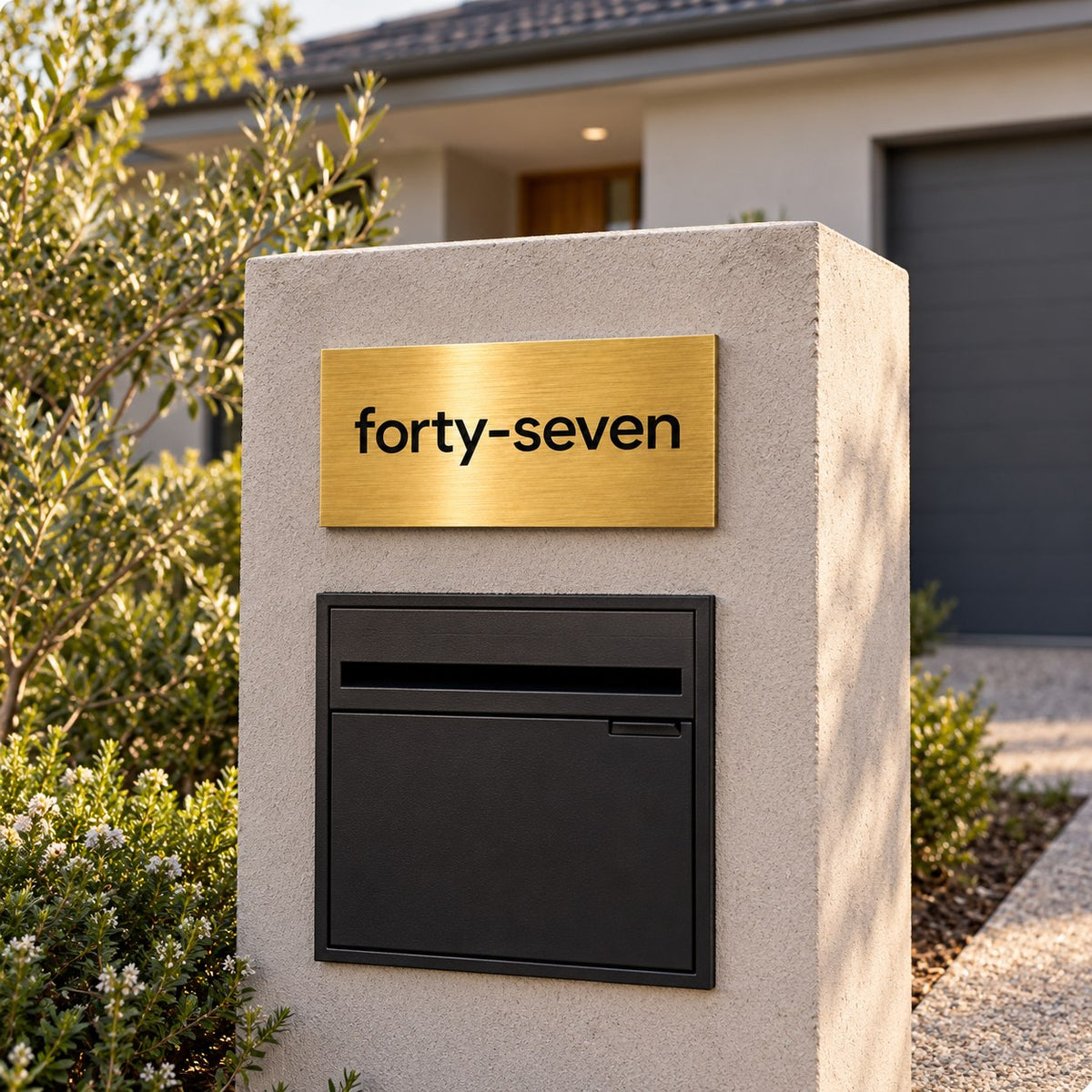 Brushed gold house number sign with a brushed metal look, The Montville, mounted on a rendered letterbox pillar above a black letterbox, the number spelled out as forty-seven in black lettering.