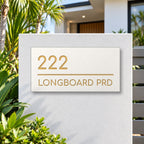 Brushed gold house number and street name sign with a brushed metal look, The Noosa, white face reading 222 Longboard Prd in gold lettering, mounted on a white rendered front pillar.