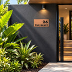 Brushed copper house number and street name sign with a brushed metal look, The Peregian, reading 26 The Bluff in black lettering, mounted on a charcoal entry wall beside the front door.