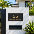Brushed gold house number and street name sign with a brushed metal look, The Peregian, black face reading 55 Oceanic Parade in gold lettering, mounted on a white rendered fence above a black letterbox.