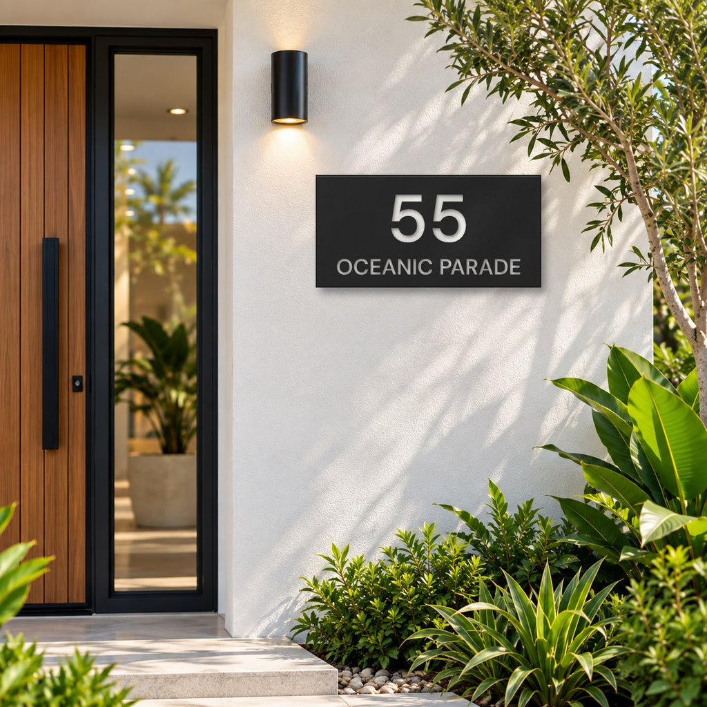 Matte black house number and street name sign, The Peregian, reading 55 Oceanic Parade in white lettering, mounted on a white rendered wall beside a timber front door.