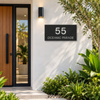 Matte black house number and street name sign, The Peregian, reading 55 Oceanic Parade in white lettering, mounted on a white rendered wall beside a timber front door.