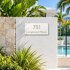 Brushed silver house number and street name sign with a brushed metal look, The Peregian, white face reading 751 Longboard Place in silver lettering, mounted on a white rendered wall beside a stone pillar and pool fence.