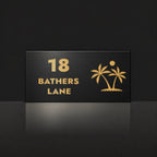 Brushed gold house number and street name sign with a brushed metal look, The Yaroomba, black face reading 18 Bathers Lane in gold lettering, with palm trees and a sun.