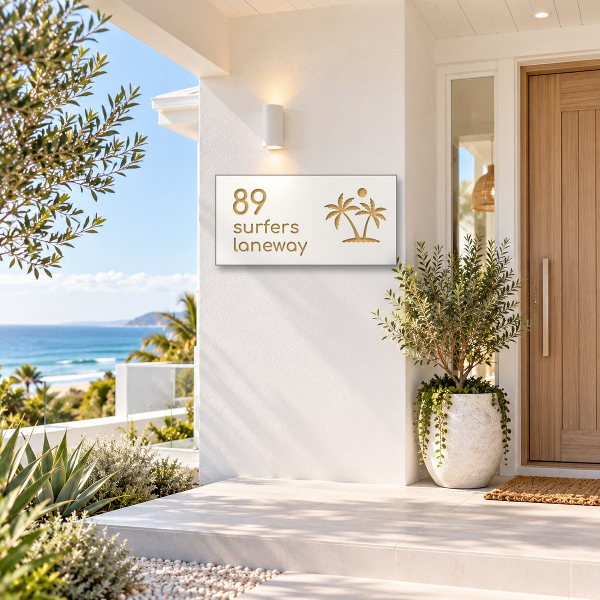 Brushed gold house number and street name sign with a brushed metal look, The Yaroomba, white face reading 89 Surfers Laneway in gold lettering, with palm trees and a sun, mounted on a white rendered wall beside a timber front door, with the ocean behind.