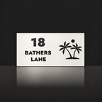 Matte white house number and street name sign, The Yaroomba, reading 18 Bathers Lane in black lettering, with palm trees and a sun.