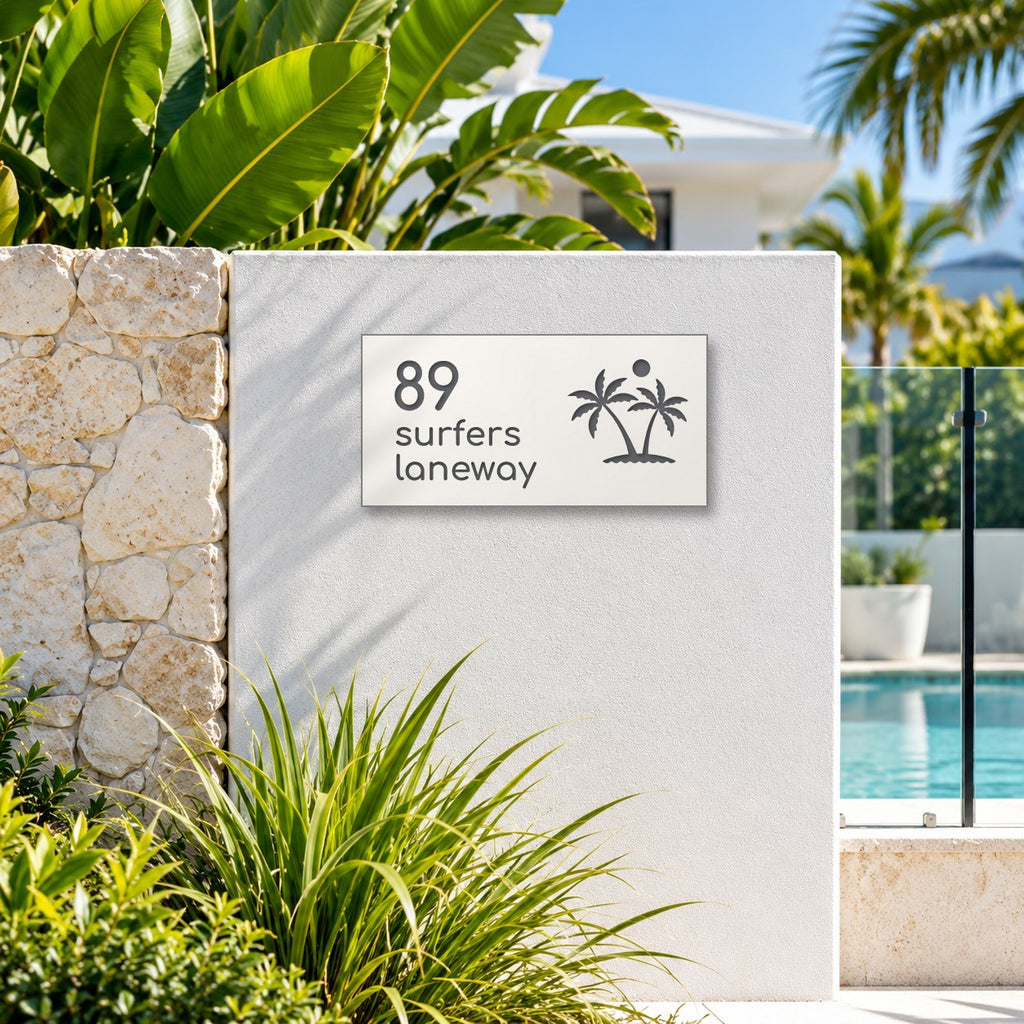Matte white house number and street name sign, The Yaroomba, reading 89 Surfers Laneway in charcoal lettering, with palm trees and a sun, mounted on a white rendered wall beside a stone pillar and pool fence.