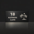 Brushed silver house number and street name sign with a brushed metal look, The Yaroomba, black face reading 18 Bathers Lane in silver lettering, with palm trees and a sun.