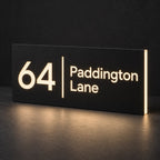 Matte black illuminated house number and street name sign, The Coolum, 64 Paddington Lane with the number, rule and street name cut through and lit from behind in warm white.