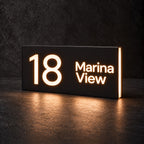 Matte black illuminated house number and street name sign, The Marcoola, 18 Marina View cut through and lit from behind in warm white.