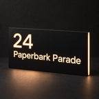 Matte black illuminated house number and street name sign, The Peregian, 24 Paperbark Parade cut through and lit from behind in warm white.