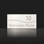Brushed silver house number and street name sign with a brushed metal look, The Bokarina, white face reading 10 Marcoola Place in silver lettering, with a wave line.