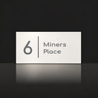 Matte white house number and street name sign, The Coolum, reading 6 Miners Place in charcoal lettering, with a dividing line.