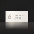 Brushed silver house number and street name sign with a brushed metal look, The Coolum, white face reading 6 Miners Place in silver lettering, with a dividing line.